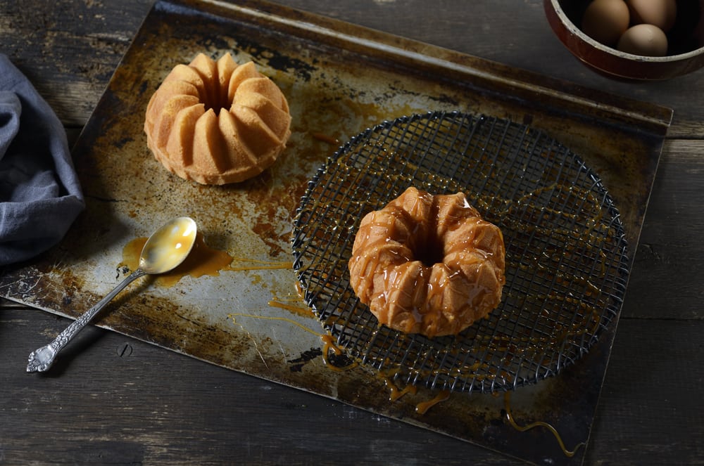 A top view of two bundt cakes with one sitting on black drain grid with caramel drizzled on it. Spoon on left with caramel in it. Bowl of eggs at top right Product Photography