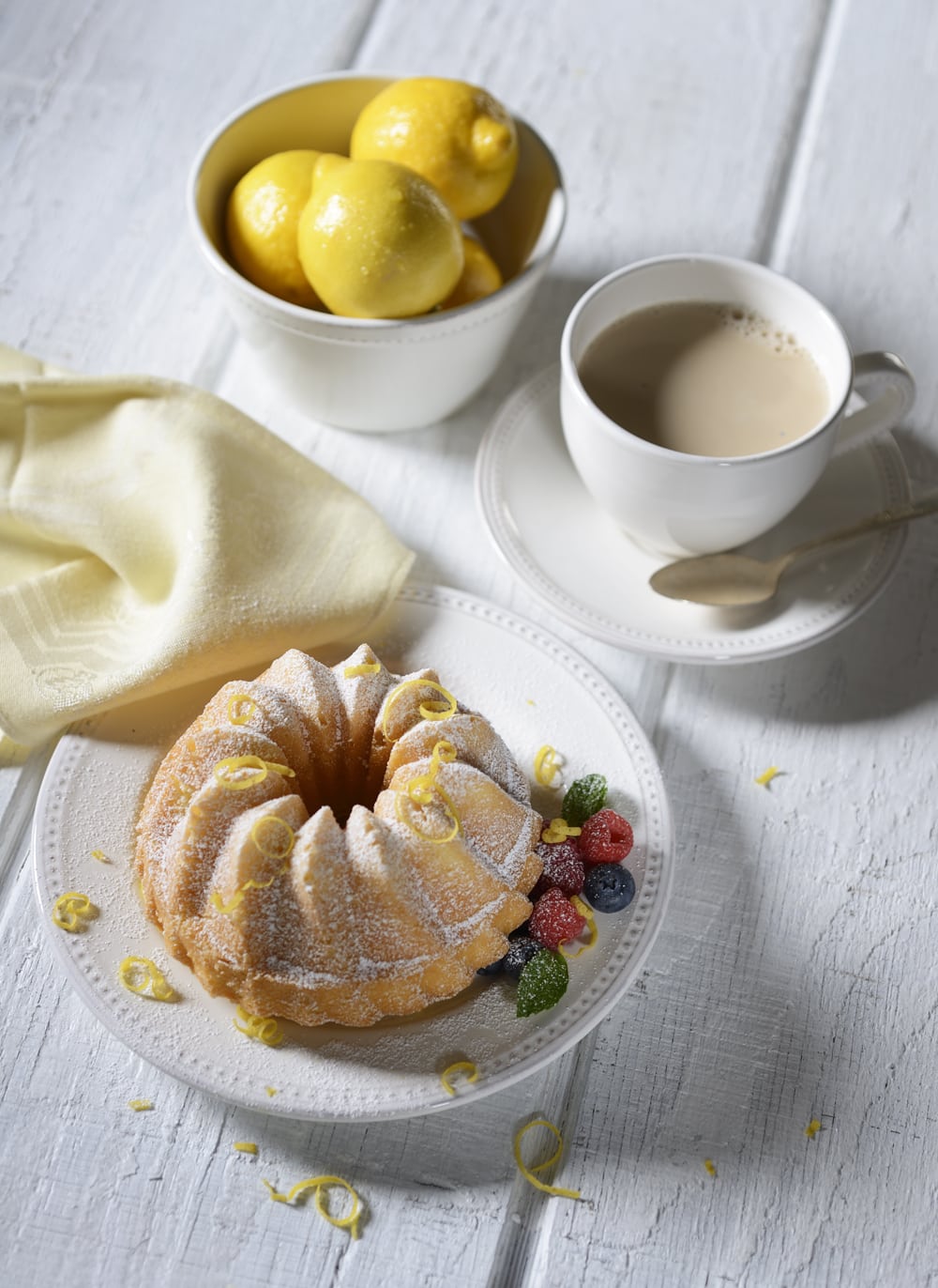 Food scene featuring a bundt cake with lemon skin shavings sprinkled on it with fruit on white plate and cup of lightened coffee and bowl of lemons all on white rustic background Product Photography