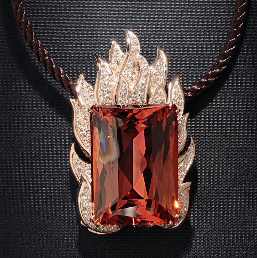 Large rare red topaz stone pendant in a rose gold flame shaped mounting with many white diamonds hanging on a brown cord. Stone features a flaw in top left corner that looks like a flame. All on black fabric background