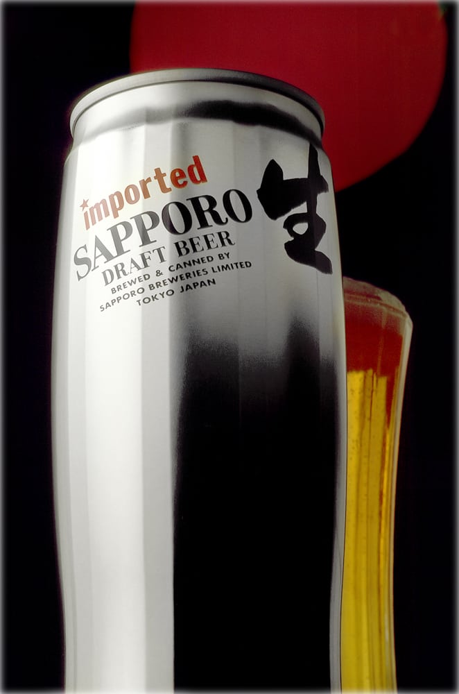 A formed beer can with the words"imported (in red) SAPPORO Draft Been..." and Japanese letters with a beer glass behind with beer and foam and a red disk on black behind Product Photography