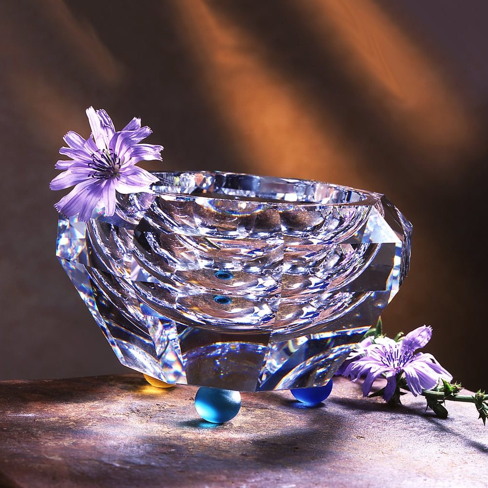 Heavy, angular crystal vase with blue and yellow glass balls for feet and purple flowers as props on textured stone background and orange light streaks behind Product Photography