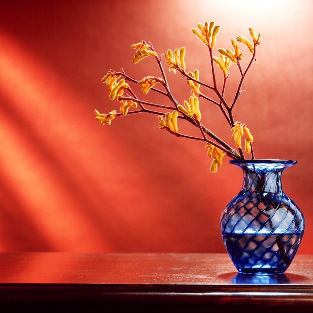 Stiking blue glass flower vase with swirl pattern holding branches with yellow buds on a brown dresser top and orange background with light streaks Product Photography